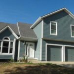 A grand house featuring an expansive driveway, set against a backdrop of trees and a sunny sky.