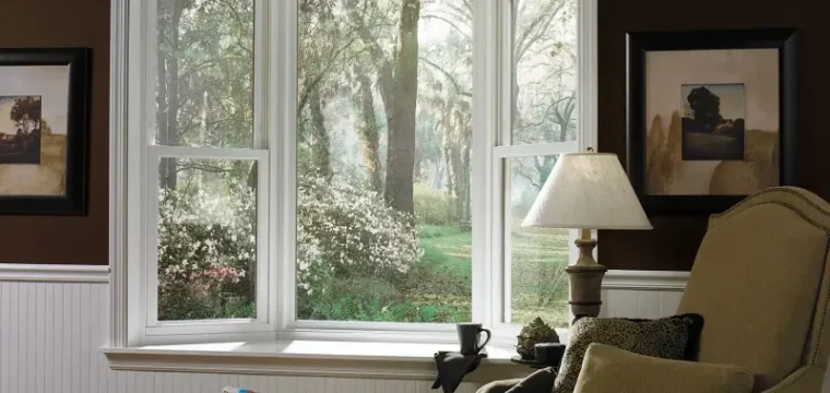 Room with an armchair and lamp by a large window. With view outside shows a sunlit forest with green trees and white flowers.